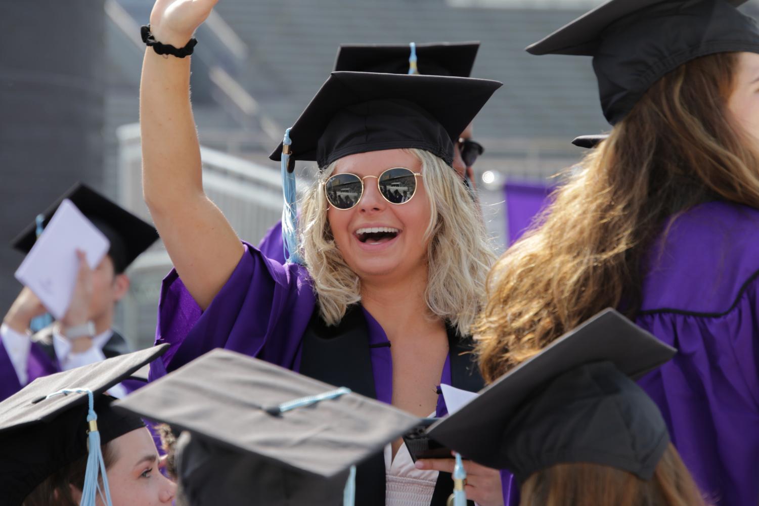 A student wearing sunglasses with a cap and gown waves to their front.