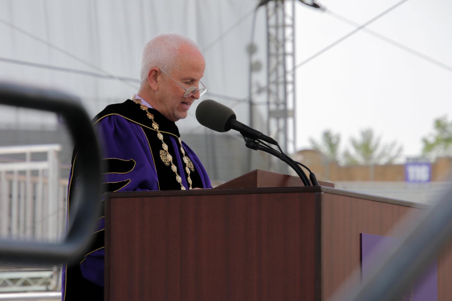 A person in a purple gown gives a speech in front of a microphone on a brown podium.