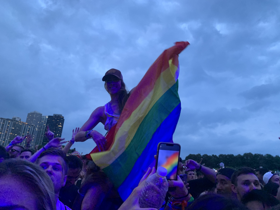 A concertgoer waves a rainbow flag while sitting on the shoulders of another participant.