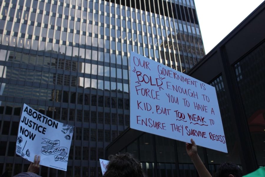 A white protest sign is held up. In red letters it reads “Our government is bold enough to force you to have a kid, but too weak to ensure they survive recess.”