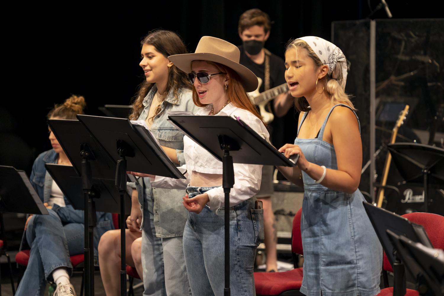Three people wearing denim sing behind music stands. The middle person wears a cowboy hat and sunglasses.