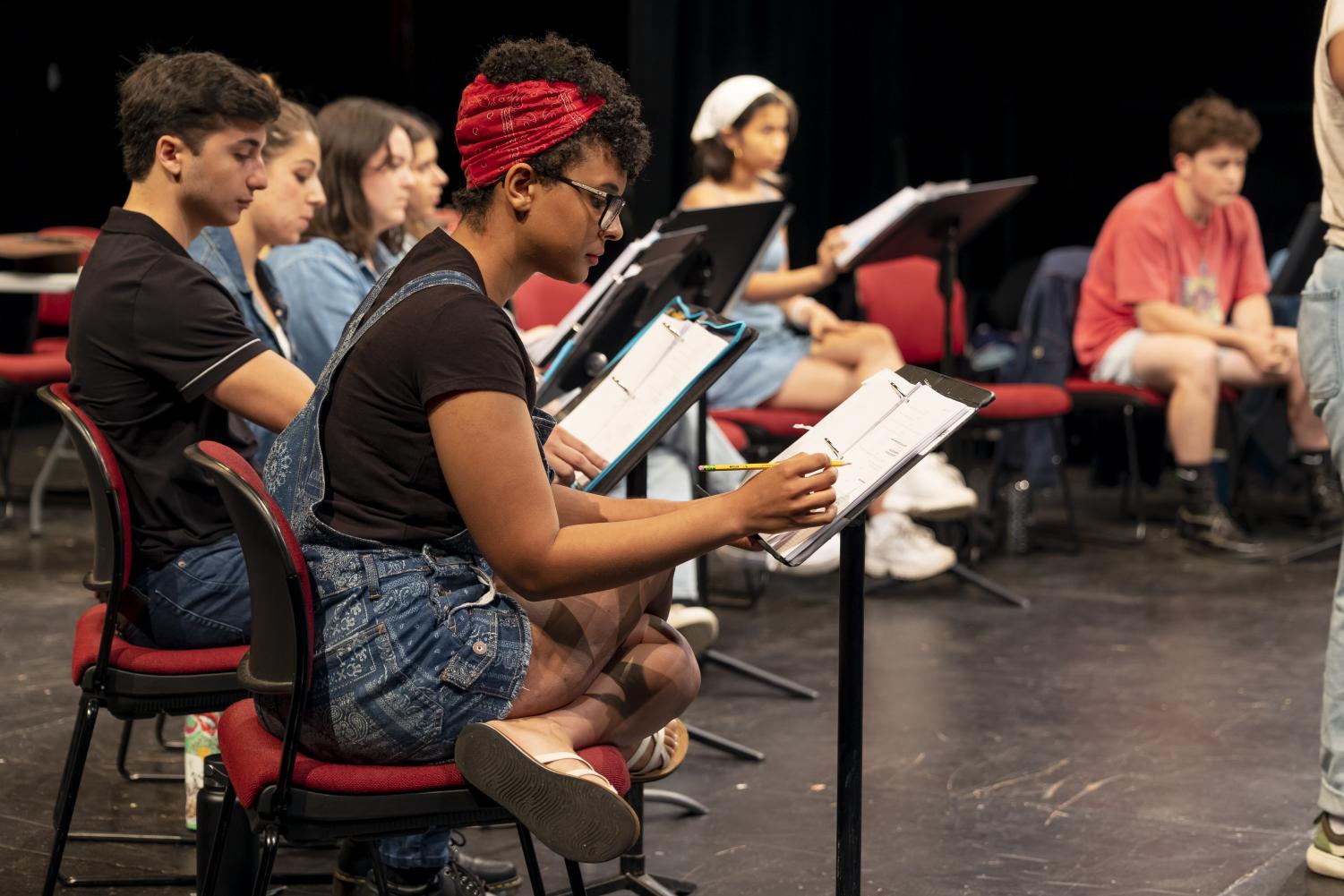 A person wearing denim overalls, a black T-shirt and a red bandana in their hair takes notes in a binder on a music stand.