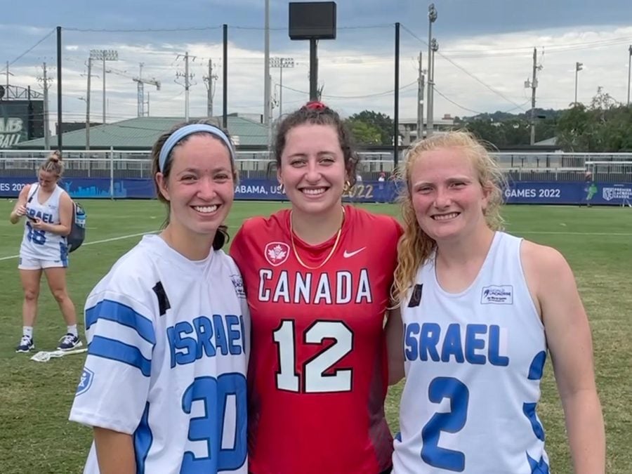 Three lacrosse players stand side by side. Two wear blue-and-white Israel jerseys and the third wears a red Canada jersey.