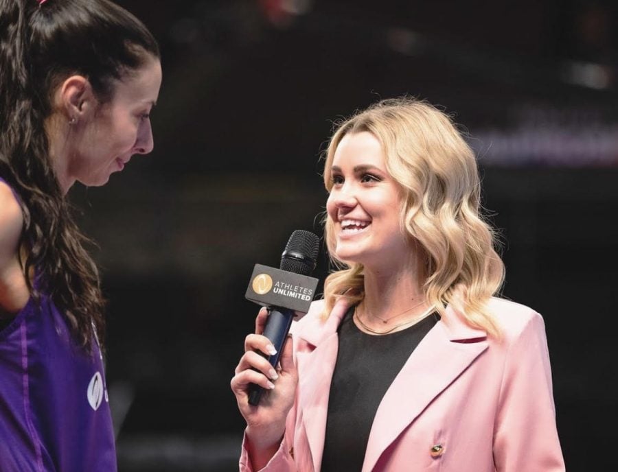 A woman+dressed+in+pink+holds+a+microphone+that+says+%E2%80%9CAAthletes+Unlimited%E2%80%9D+and talks+to+a+athlete+dressed+in+a+purple+jersey.