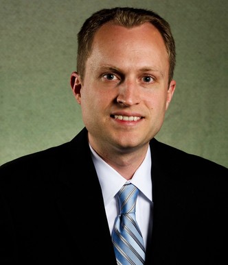 Luke Stowe poses for a portrait. He is wearing a Black suit, his face is head-on and the background is green.