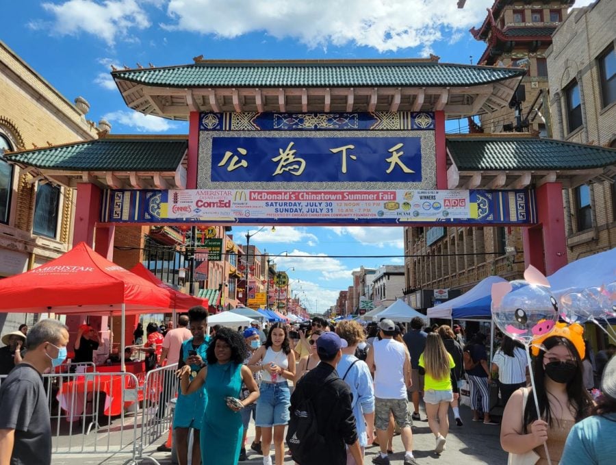 The Chinatown gate with a Summer Fair poster is surrounded by people walking between tent booths.