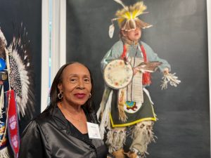 A woman with large earrings, wearing a black leather jacket stands in front of a life size photograph of a child dressed in traditional Native American ceremonial clothing.