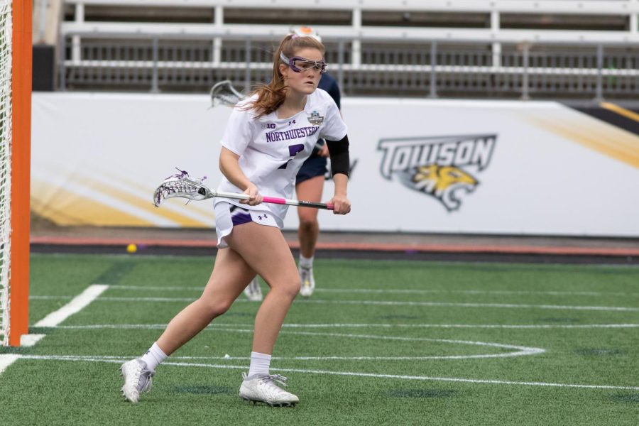 A lacrosse player wearing purple and white runs downfield holding a stick.