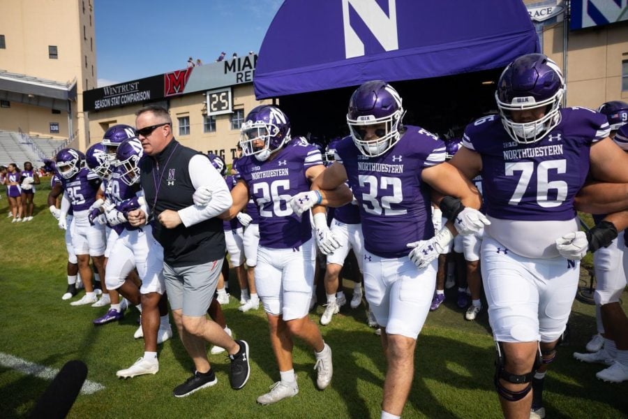 Football players in purple uniforms and a coach in a black shirt walk onto a football field.