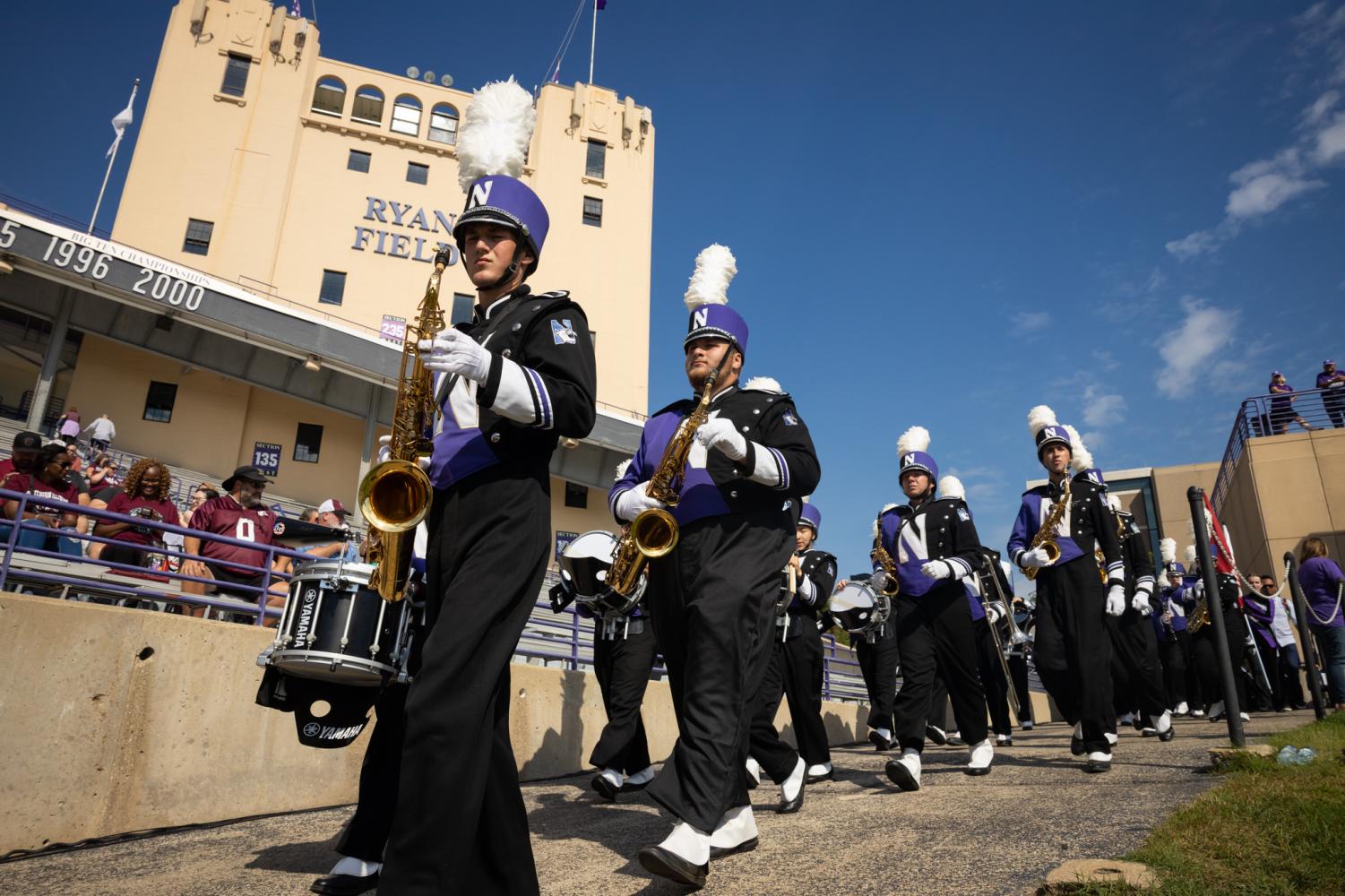 A marching band dressed in black and purple walks down a ramp and onto a football field. 