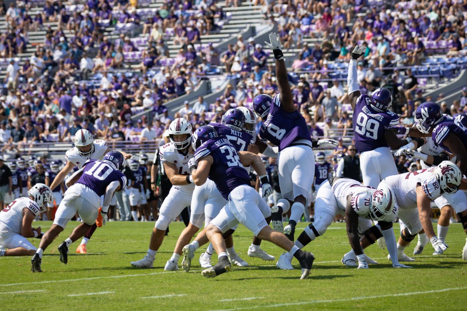 Football players in purple jerseys jump to try to block a field goal. 