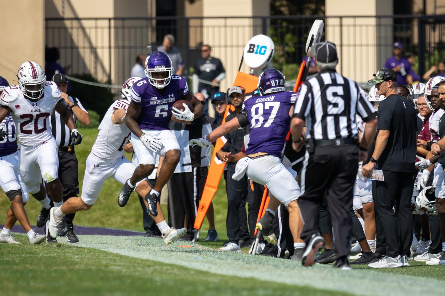 A football player in a purple jersey jumps onto the sidelines to try to avoid a tackle by a player in a white jersey.