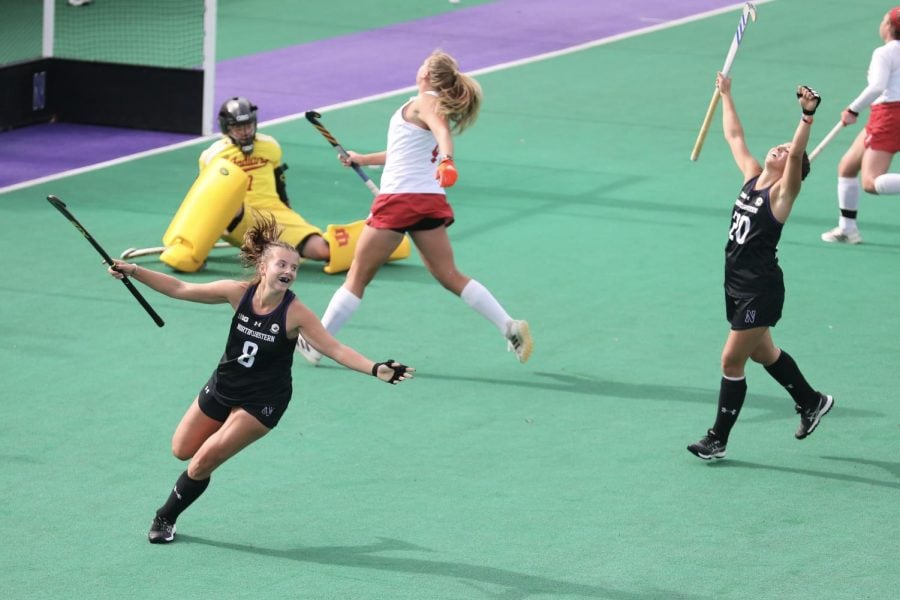 A field hockey player in a black jersey raises her arms and celebrates after scoring.