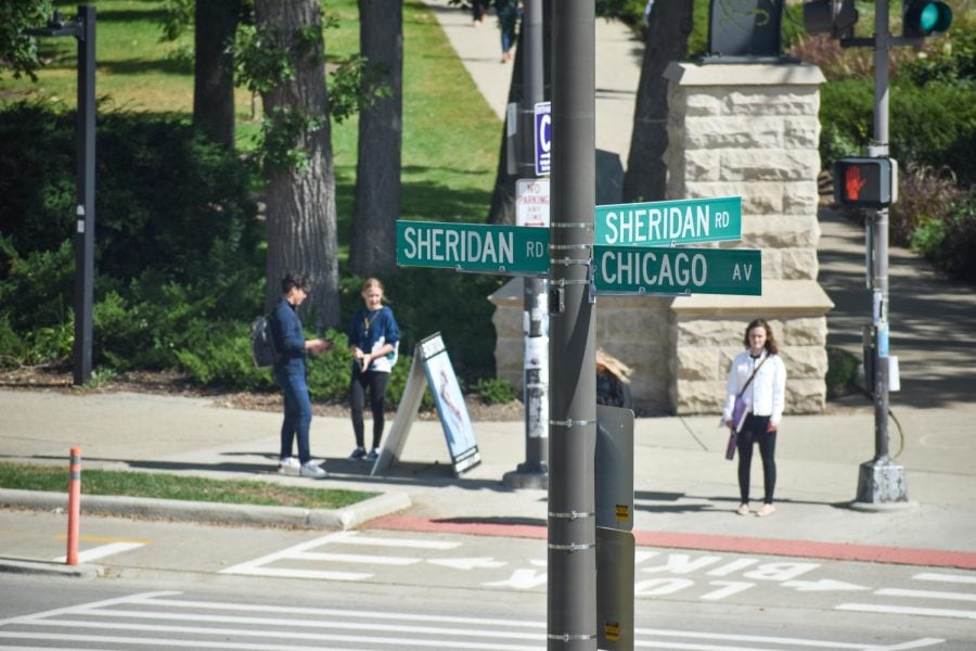 Green street signs reading “Sheridan Rd” and “Chicago Av” with people standing on a sidewalk corner in the background.