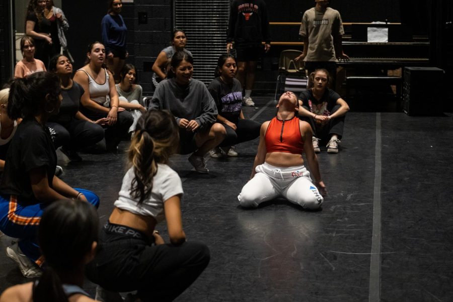 A dancer wearing a red top and sweatpants kneels and tilts their head up, surrounded by a crowd of people sitting and crouching in a room with black floors and walls.