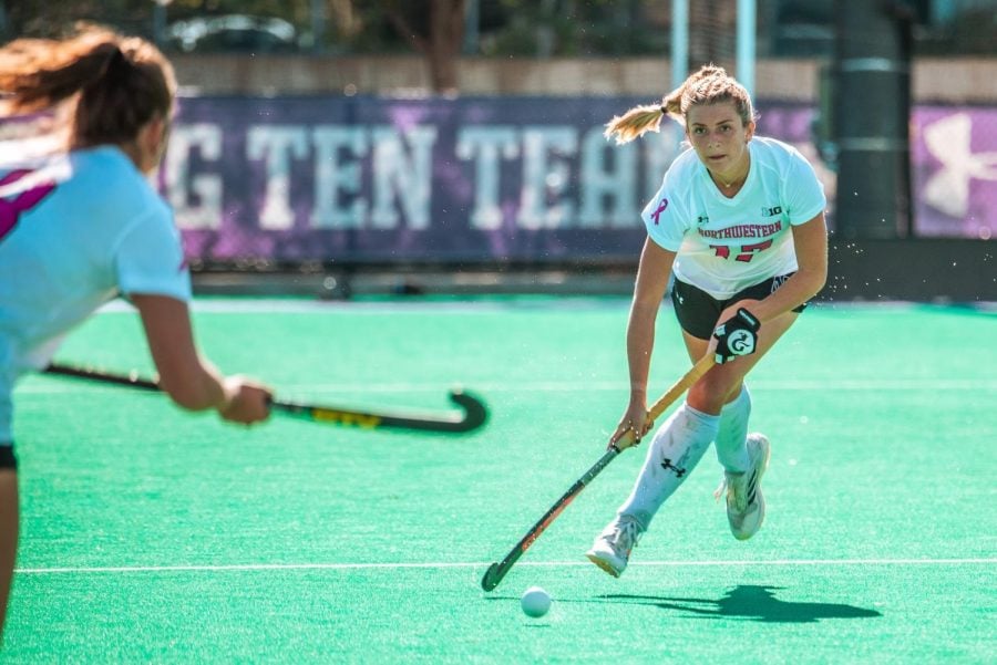 A field hockey player in a white and pink jersey with a breast cancer awareness ribbon on it dribbles the ball down field.