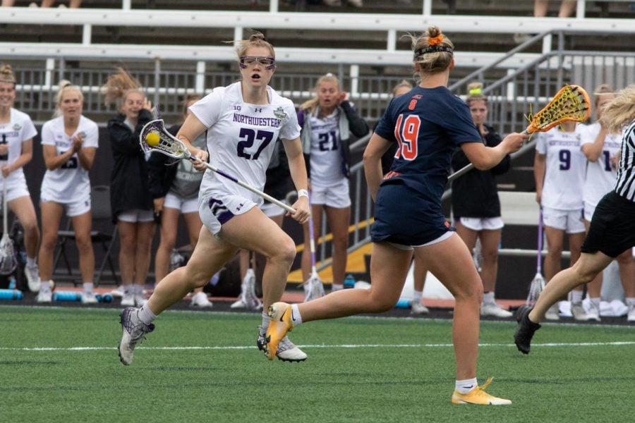A girl with brown hair runs down a field holding a lacrosse stick as a player defends her.