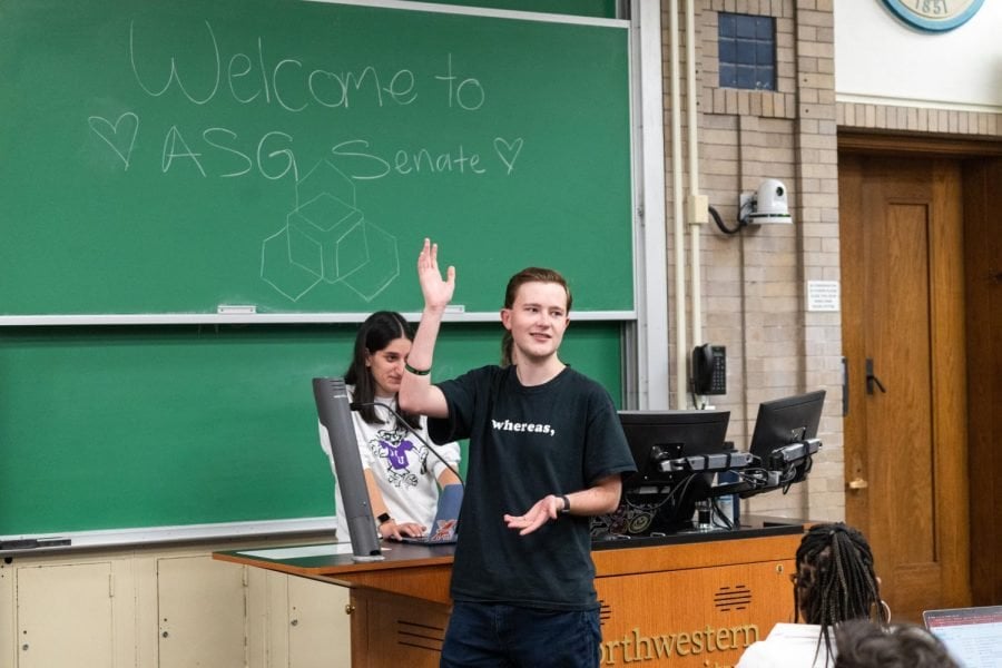 A person in a black shirt waves to a crowd while standing in front of two other people and a green chalkboard which reads, “Welcome to ASG Senate.”