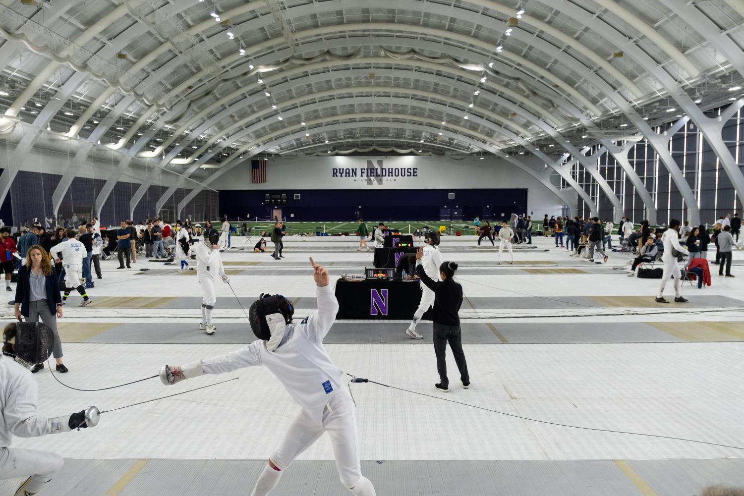 Fencers, referees and onlookers mill about in a large room with a curved roof.