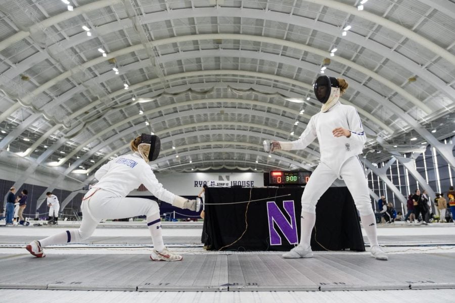 One fencer crouches to hit another fencer in a large room with a curved roof.