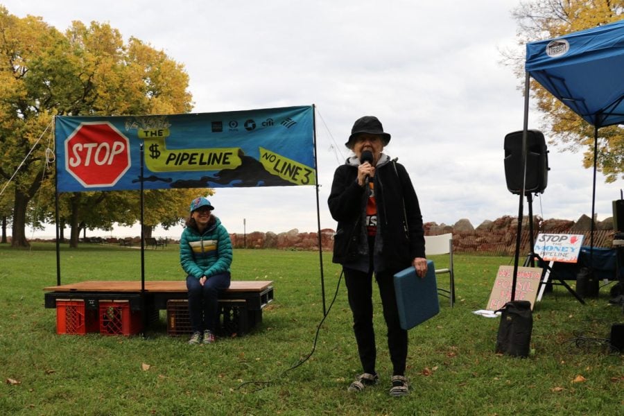 An Evanston resident dressed in all black holds a microphone and speaks to a crowd. In the background, rally emcee Jessy Bradish sits on a small wooden stage. A banner reading “stop the money pipeline” and depicting an oil leak hangs above the stage.