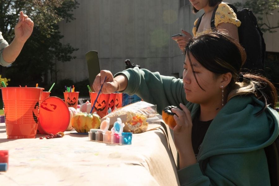 A student kneels next to an outdoor table at the Fall Festi-Ful, painting a pumpkin surrounded by orange plastic cups.