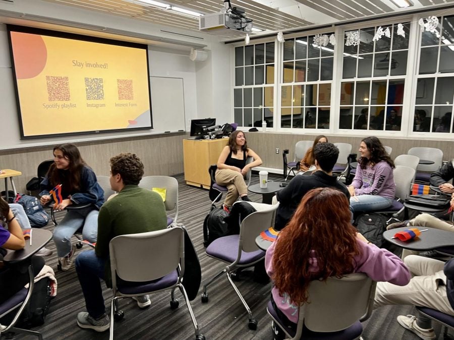 Students gather in a classroom sitting in chairs, with a window in the background on one side and a projection screen and whiteboard on the other.