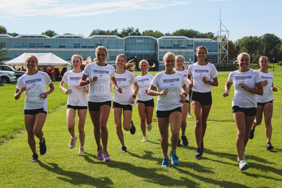 Ten athletes in white shirts and black shorts jog forward while smiling.