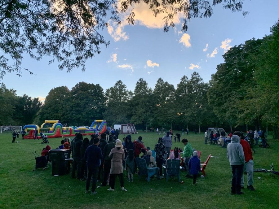 People gather around picnic tables in front of bouncy castles at sunset. They are in a grassy park surrounded by trees.