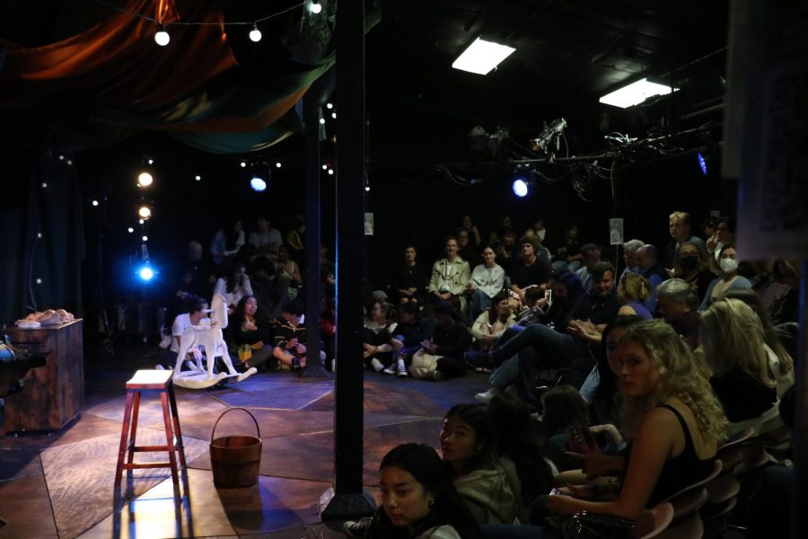 People sit on the floor and in chairs around a stage with props on it, waiting for the show to start.