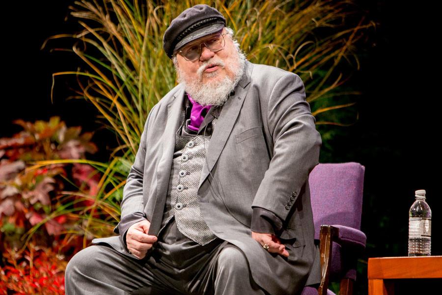 George R.R. Martin sits on a purple chair with a colorful background.