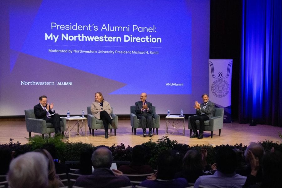 Four people sit on a stage clapping with “President’s Alumni Panel: My Northwestern Direction” projected on a screen behind them.
