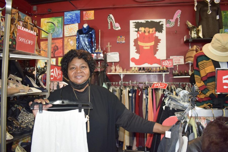 Mary Toussaint holds black hanger with white slip in front of racks of clothing and a red wall with paintings on it.