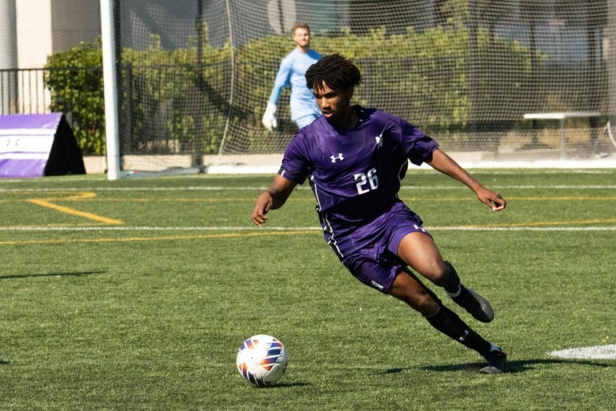 A soccer player in a purple jersey and purple shorts dribbles the ball.