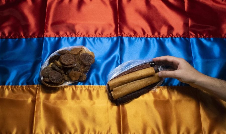 A hand of a person reaches to grab dry fruits. The fruits are placed on a red, dark blue and yellow Armenian flag, serving as a tablecloth.