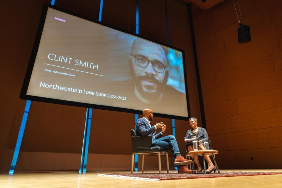 A screen with a man’s face hangs down from the ceiling with the words “Clint Smith” next to it.