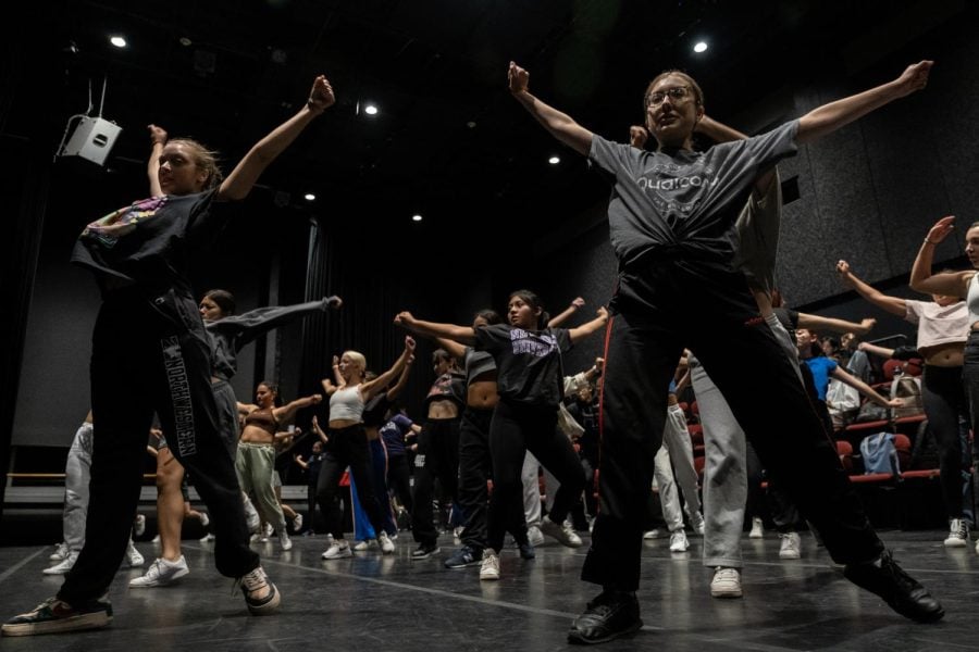 People stand with their arms raised wearing gray shirts at a dance performance.