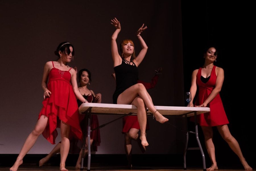 Four dancers in red costumes gather around a dancer in a black dress in the center.