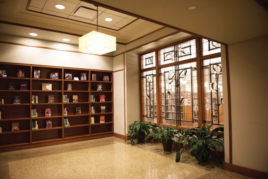 Inside Evanston Public Library, 1703 Orrington Ave., there are three dark brown bookshelves with books dispersed on them. Light shines through three glass windows with plants underneath them.