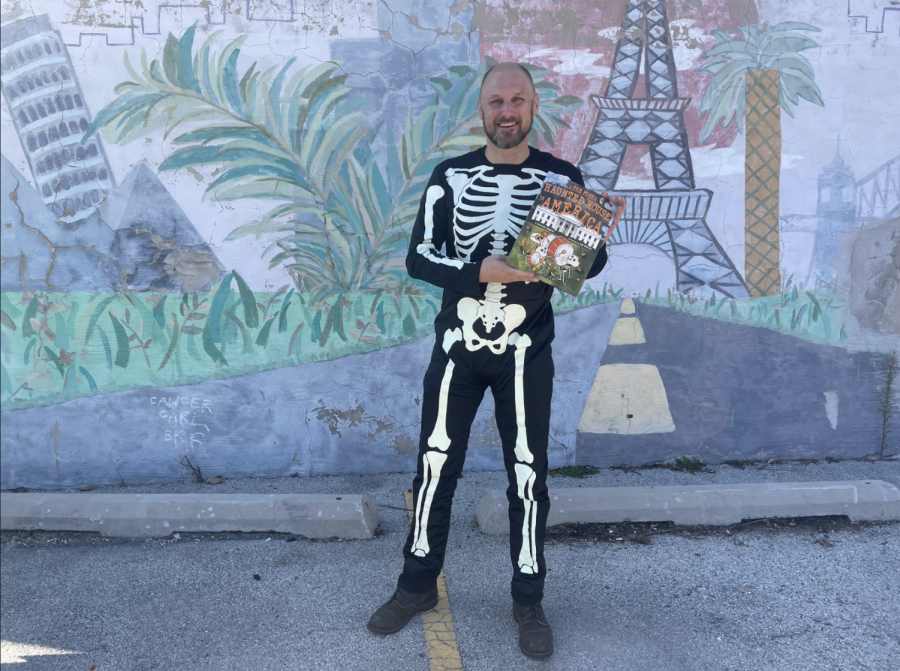 A man in a skeleton costume holds up a book in front of a colorful wall.