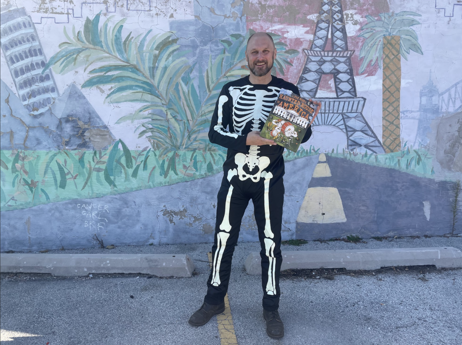 A+man+in+a+skeleton+costume+holds+up+a+book+in+front+of+a+colorful+wall.