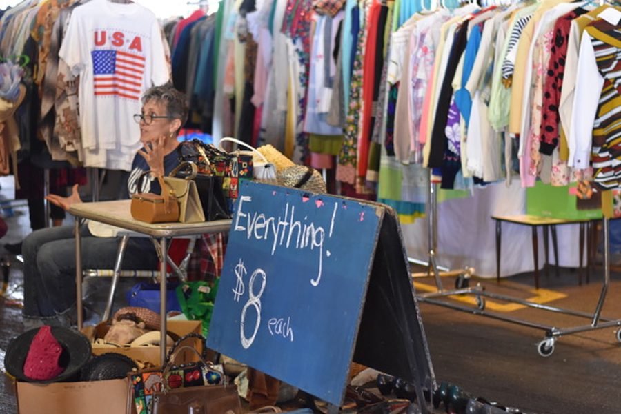A sign reads “Everything $8” in front of a vendor’s shop at a Vintage Garage flea market. A vendor is sitting in a chair talking to customers with rows of clothes and tables with hand bags surrounding her.