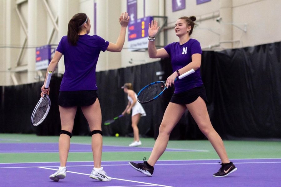 Two women in purple shirts and black shorts holding tennis rackets high five on a tennis court.