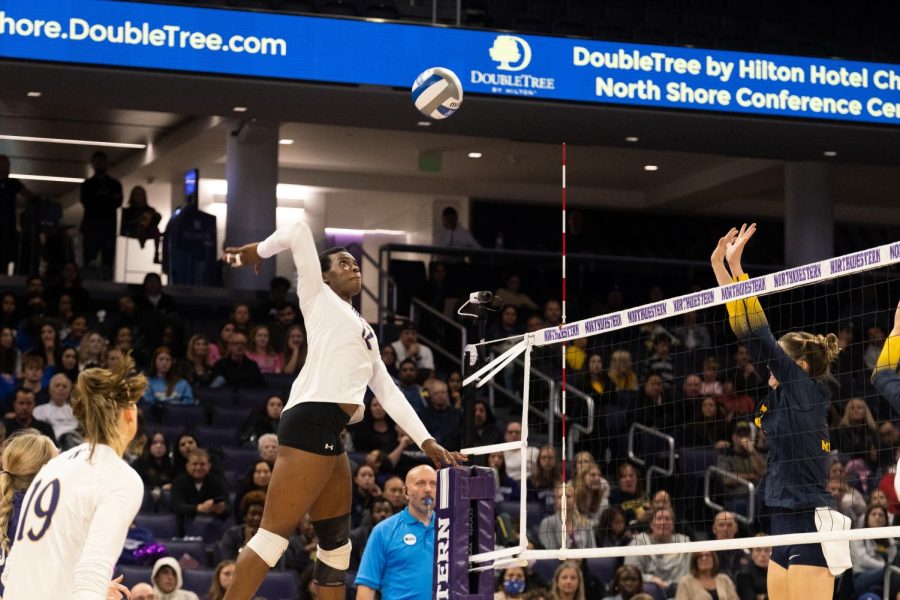A woman wearing a purple-and-white jersey reaches to hit a volleyball.