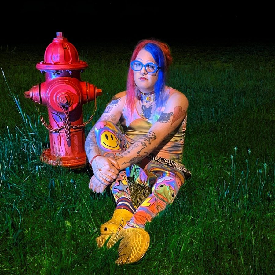 A person with blue and pink hair wearing colorful patterned clothing and yellow crocs sitting in green grass next to a red fire hydrant.