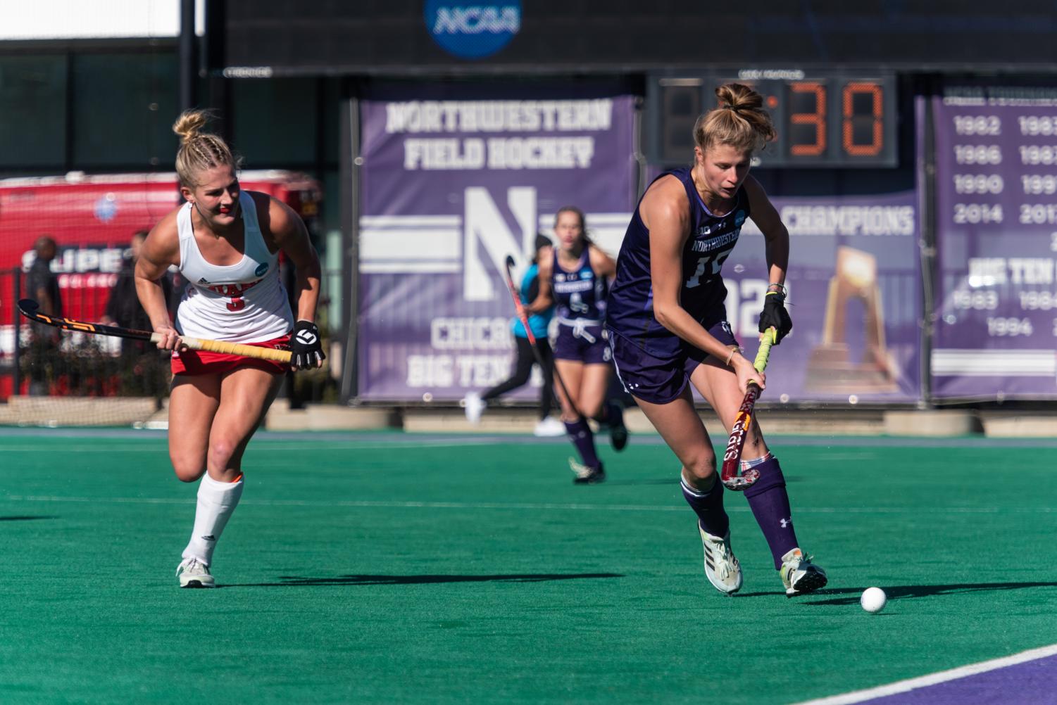 A field hockey player in a purple jersey runs while keeping the ball away from a player in a white jersey.