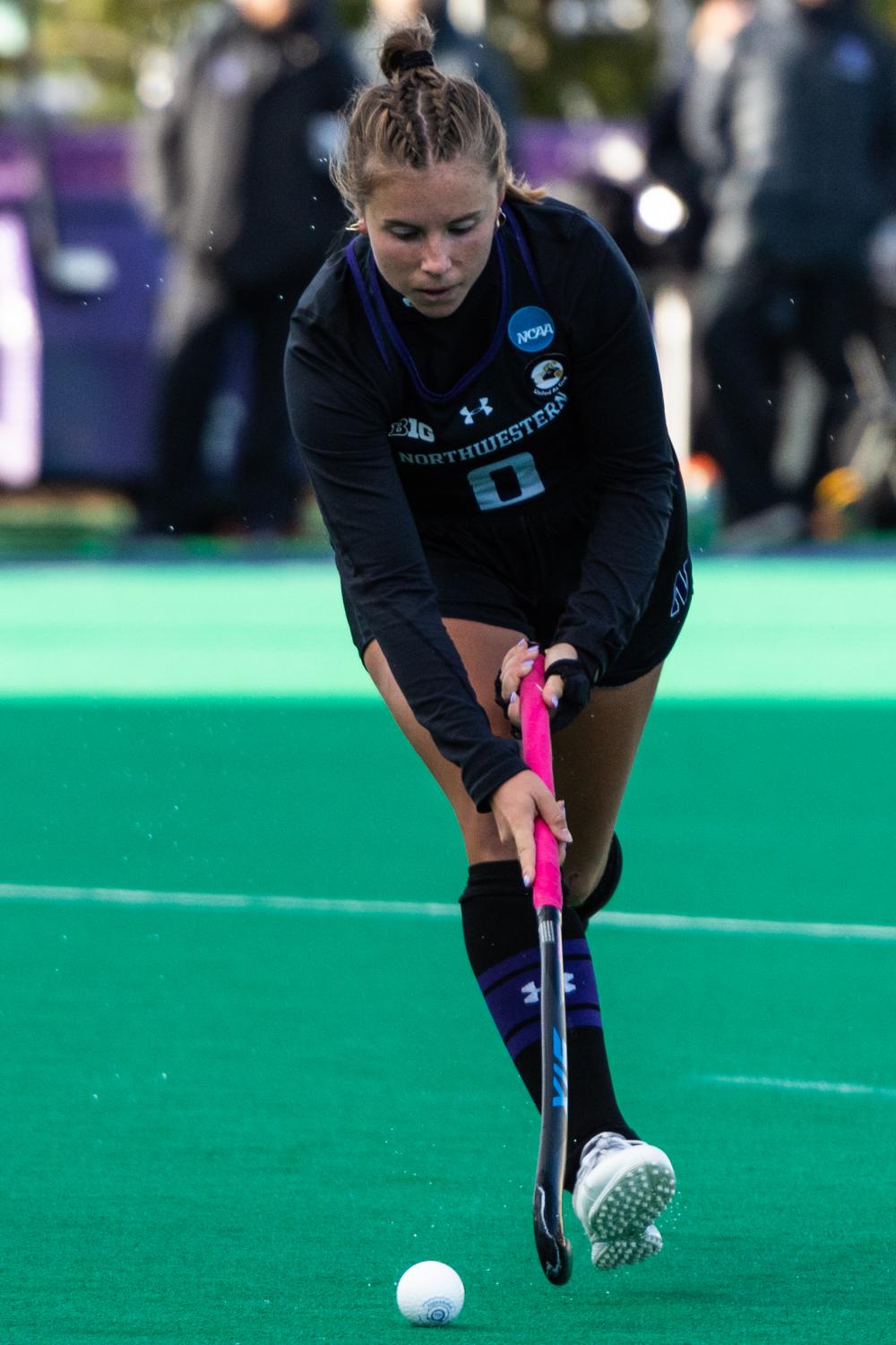 An athlete in a black jersey holds a field hockey stick forward while tracking a white ball.