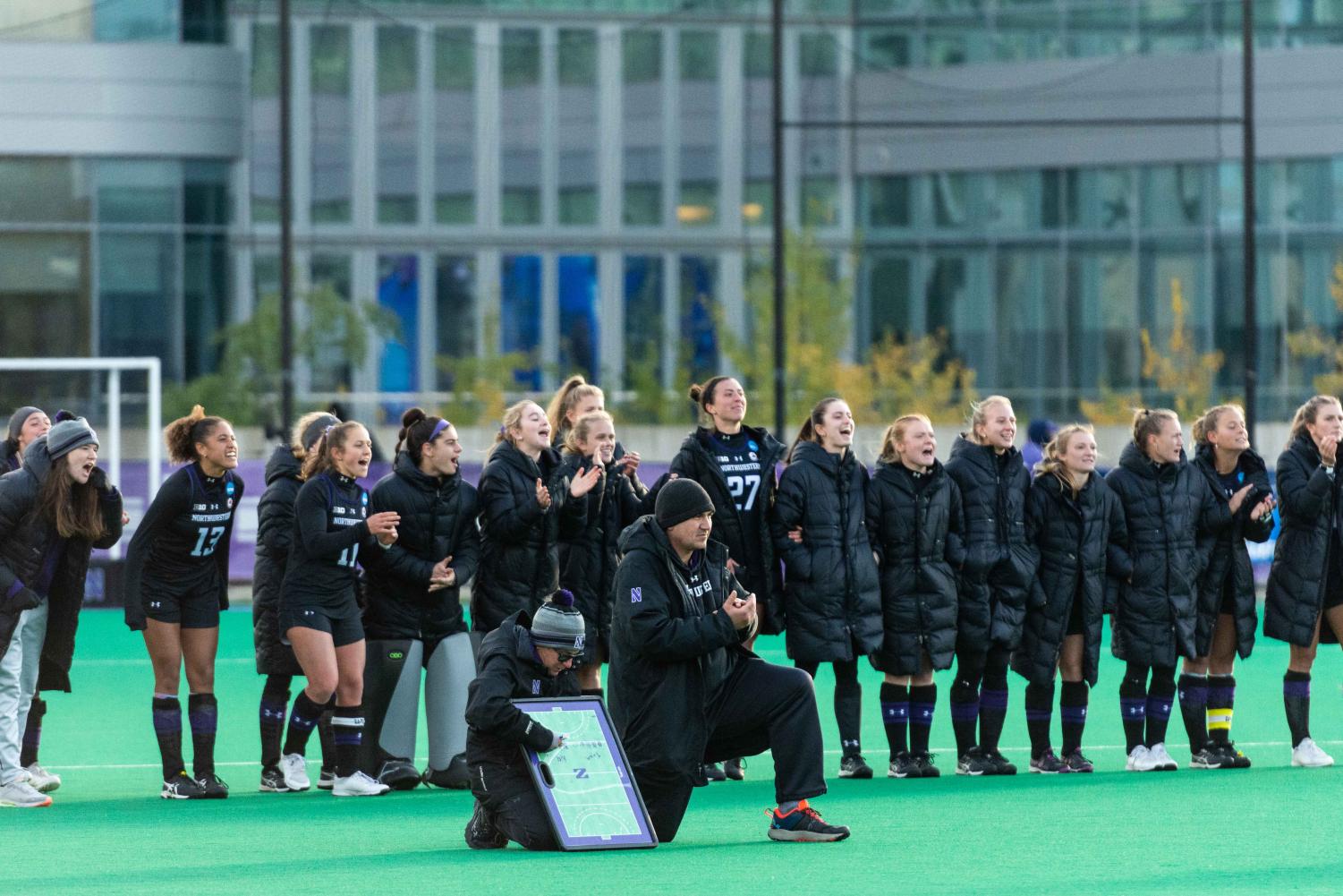 Two coaches, one holding a whiteboard, kneel in front of a row of athletes in black jerseys and black coats.