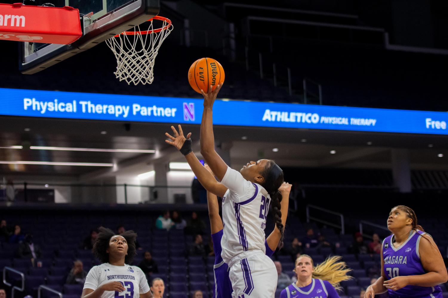 A basketball player in white jumps to bring the basketball closer to the hoop as they attempt to shoot while other players in purple and white watch.
