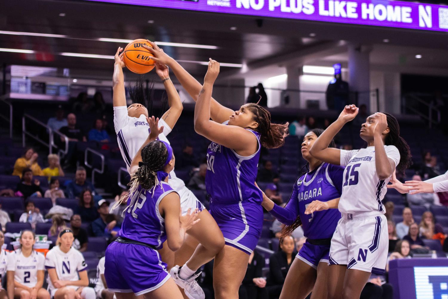 A basketball player in white catches the basketball as other players in purple and white try to reach for it.